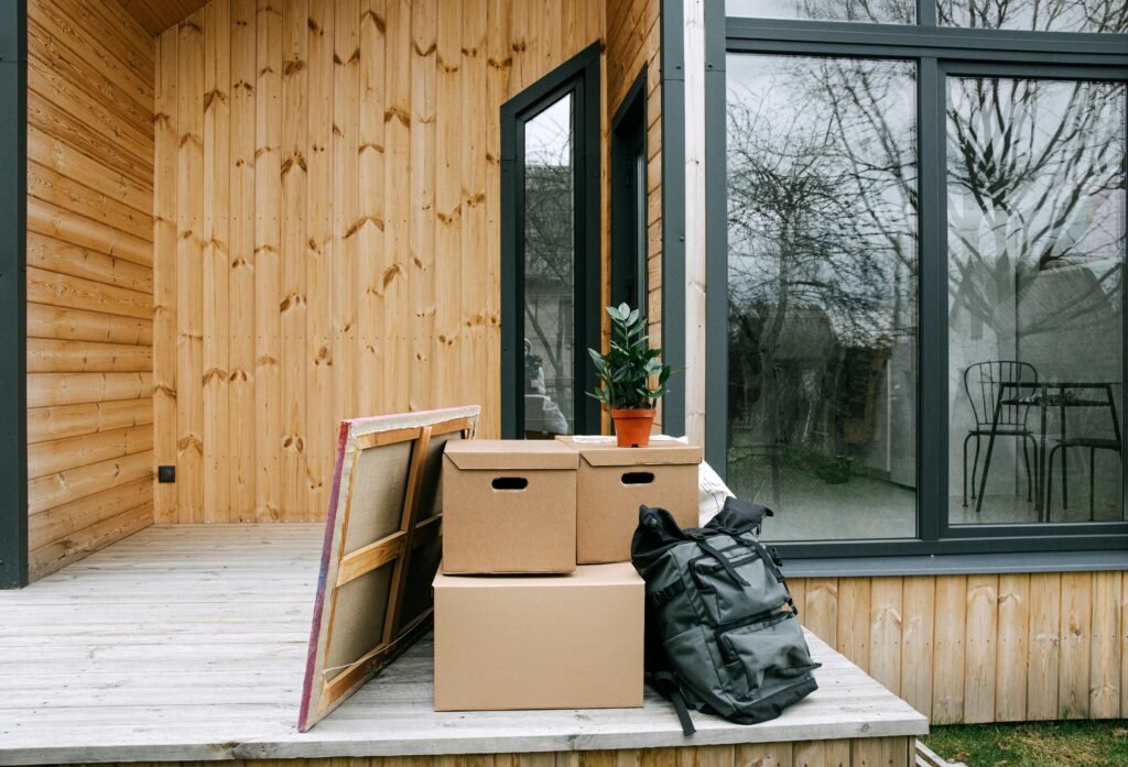 Wooden porch with cardboard boxes and potted plant next to glass windows.